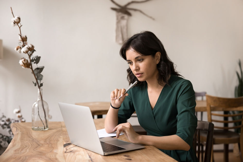 A woman is seated at a table in front of a laptop computer. She is holding a pen, looking at the screen, and thinking.