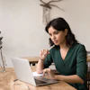 A woman is seated at a table in front of a laptop computer. She is holding a pen, looking at the screen, and thinking.
