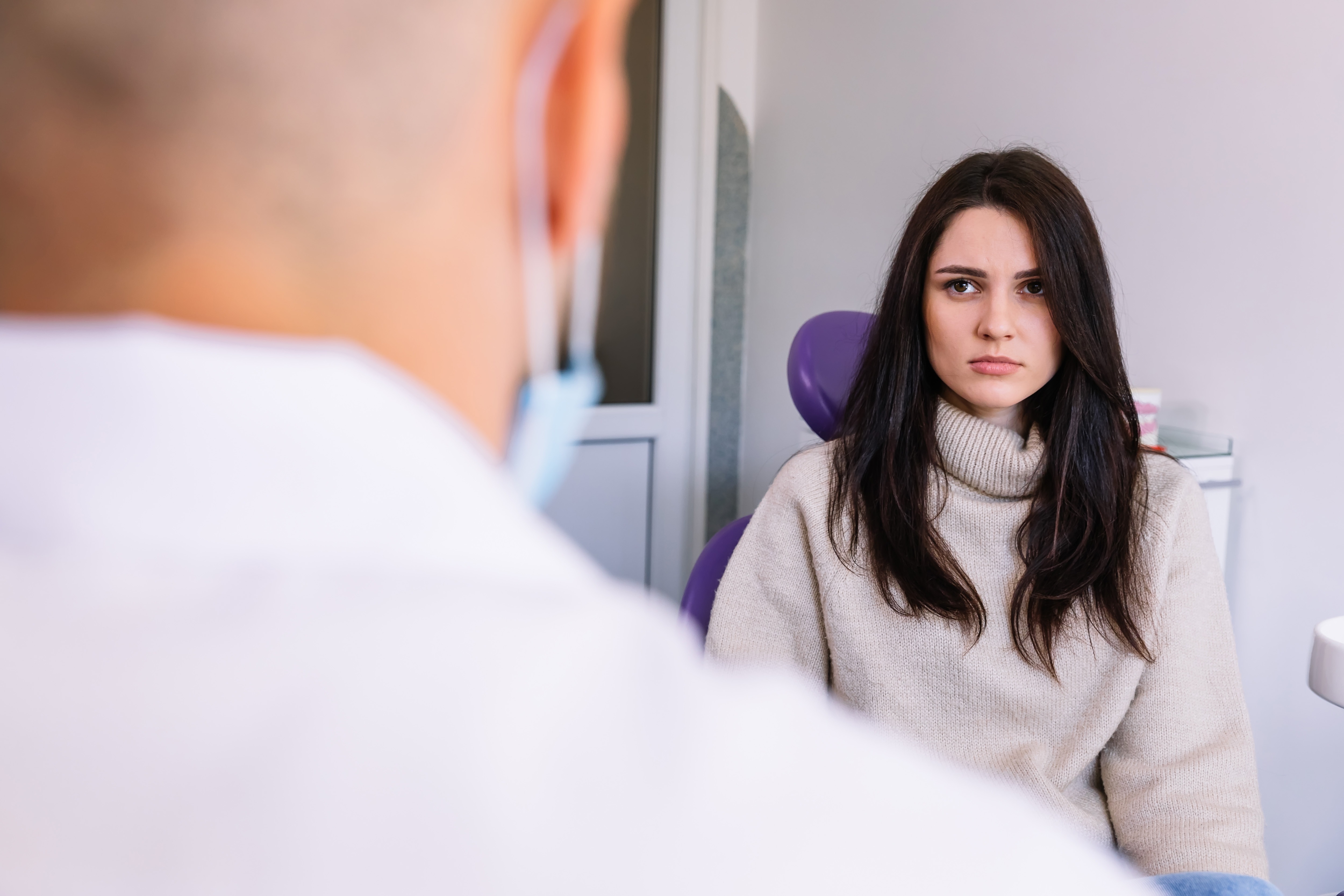 A woman sitting in a dental chair has a serious expression on her face while a dentist, who is in the foreground, talks to her.