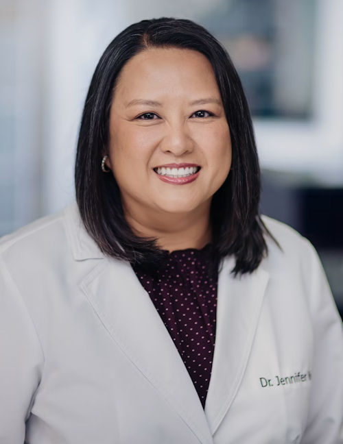 Professional headshot of a smiling woman wearing a white medical coat and dark patterned top